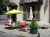 Барбизон - Фасад дома и тротуара украшают куст в горшке, столы, стулья и зонтик