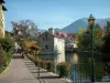 Annecy - Banco de floração, postes de iluminação, árvores, canal de Thiou, casas na cidade velha à beira da água e montanhas ao fundo
