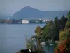 Lac d'Annecy - Arbres aux couleurs de l'automne, maisons au bord du lac, bâtiments de la ville d'Annecy et colline en arrière-plan