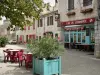 Lauzerte - Arbuste fleuri en pot, terrasse de café et façades de maisons de la place des Cornières
