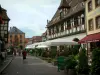 Obernai - Calle pavimentada, con casas de madera y ventanas adornadas con flores (geranios), la terraza del restaurante y el ayuntamiento (alcaldía) en el fondo