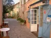 Ramatuelle - Narrow street of the village, tables and chairs of a bar, plants and flowers in jars, houses with colourful facades