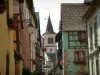 Riquewihr - Houses with colourful facades decorated with forged iron shop signs and flower-bedecked windows (geranium), the church bell tower in background