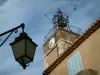 Tourtour - Farola en primer plano, la torre coronada por un campanario de hierro forjado y la casa de verano