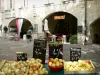 Uzès - Place aux Herbes: puesto de frutas y verduras del mercado en el primer plano, restaurante con terraza, tienda y hogar de arcadas (arcos)